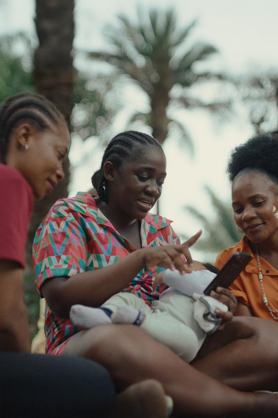 A woman sits outside with her baby in her lap and shows her two friends something on her phone in her hand.